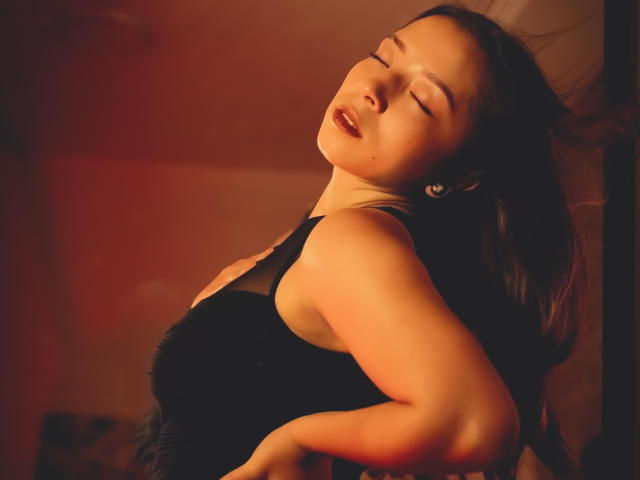 NatashaBlank with eyes closed, head tilted back, black dress, soft red light, arms slightly raised, dark hair, earring, sensual expression, her tits and ass subtly visible.