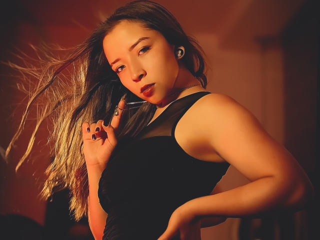NatashaBlank with long brown hair, wearing a black sleeveless top, showing off her tits, holding her hair, red lighting, black nails, earbuds, and a ring.