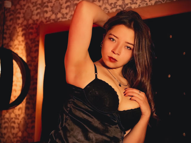NatashaBlank with dark brown hair, wearing a black velvet dress, exposing her tits with a raised arm, looking seductively at the camera, in a warmly lit room.