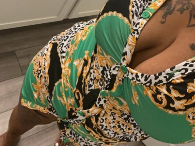 El escote de JessyXBabe69 es muy visible en un vestido de estampado de leopardo en verde, negro y dorado, que muestra sus grandes senos redondos y un tatuaje en su hombro derecho. Está inclinada hacia adelante..