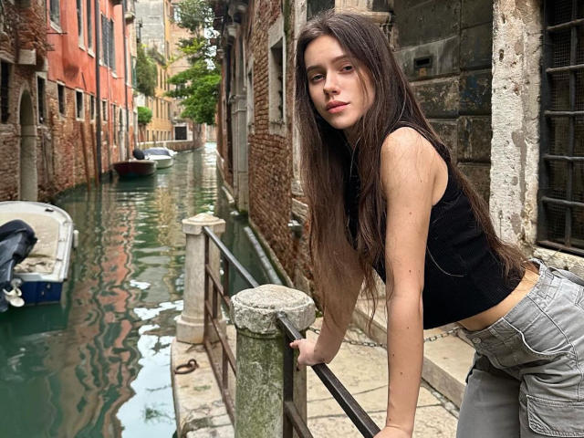 MoonSerenade with long brown hair, black sleeveless top, and gray pants, leaning on a rail by a green canal, looking at the camera with a serious expression.