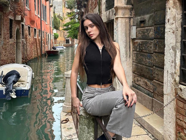 MoonSerenade with long brown hair, black sleeveless top, gray pants, sitting on a wooden post, looking serious, next to a green canal with a small boat.