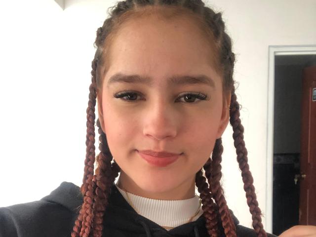 Melody Mansson tiene la piel de color marrón claro, una nariz pequeña y labios rosados y gruesos. Lleva el cabello rojo trenzado, una sudadera con capucha negra y una camiseta blanca con textura. Está sonriendo suavemente..