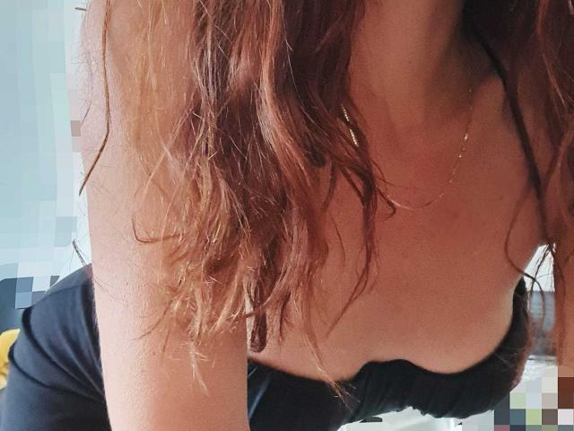 Varela69 met golvend roodbruin haar, leunt voorover en laat haar grote borsten zien in een zwarte top zonder bandjes, draagt een gouden ketting, het bovenlichaam en de arm zijn zichtbaar..