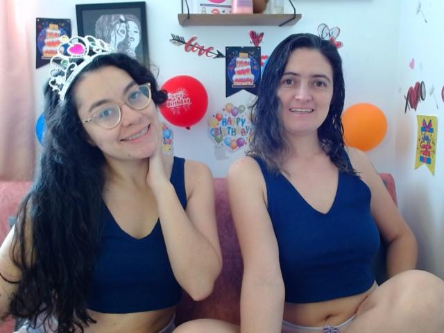 LunaVenus with long black hair and glasses, wearing a silver tiara, and LunaVenus with wet black hair, both in blue sports bras, smiling, sitting on a pink couch.