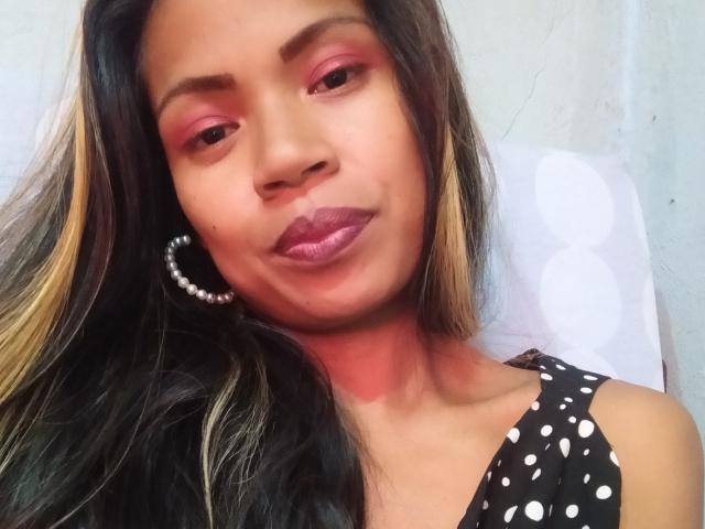 Cupcake69 has dark brown skin, pink eyeshadow, shiny purple lipstick, black and blonde hair, hoop earring, and wears a black polka dot top, showing a confident smile.