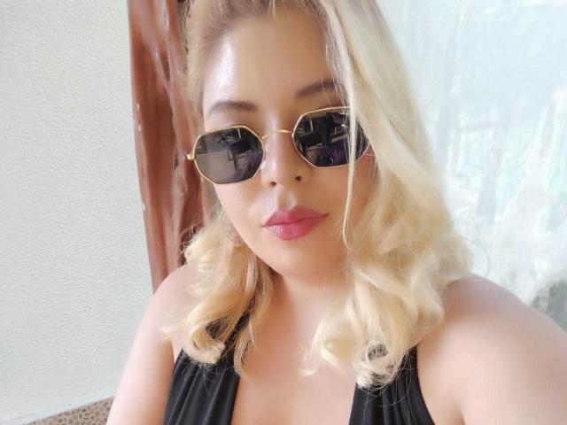 Blonde MilenaDutty with wavy hair, wearing black sunglasses, and a black halter top, has pink lipstick and a serious expression. Her tits are small and her ass is unseen.