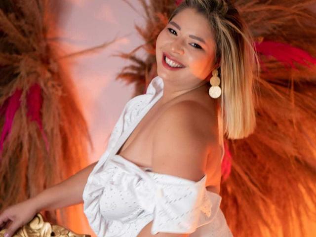 MilenaDutty with blonde hair, smiling, wearing a white off-shoulder top, exposing her tits, round earrings, and golden chair, in a warm, orange-lit setting.