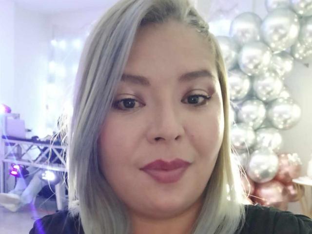 MilenaDutty with pale skin, light gray bob, and pink lipstick, pouting her full lips, wearing black top, surrounded by silver balloons in a softly lit room.