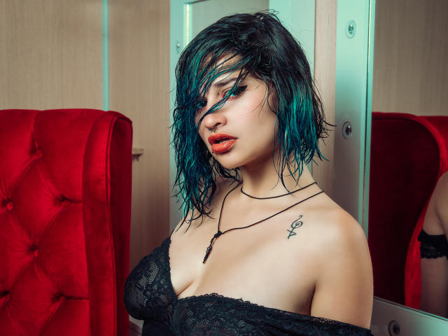 MaiaThunder, con el cabello mojado teñido de color turquesa, vestida con una blusa negra de encaje con un hombro descubierto, mostrando sus pechos, labios rojos, aro en la nariz, tatuaje en el hombro izquierdo, de pie frente a una silla roja..