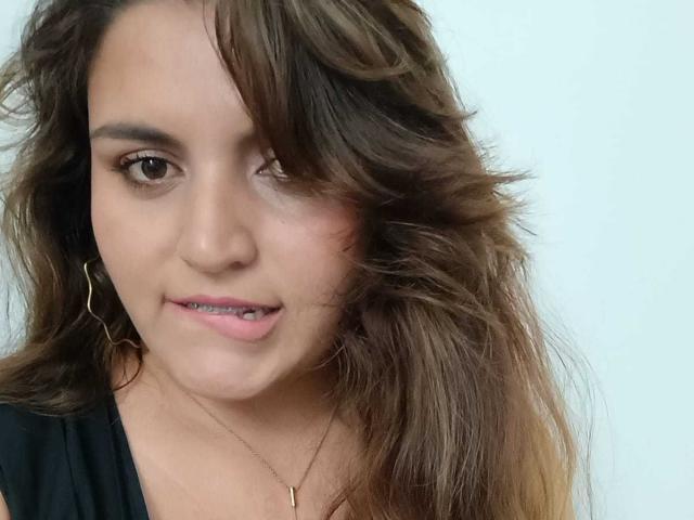 SharaySexy has wavy brown hair, light skin, and is sticking her tongue out slightly. She's wearing a black top and gold hoop earrings. Her eyes are brown and she's smiling.