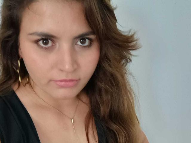 SharaySexy has wavy brown hair, brown eyes, and pink lips, wearing a black top and gold hoop earrings with a thin necklace, looking slightly to the side.