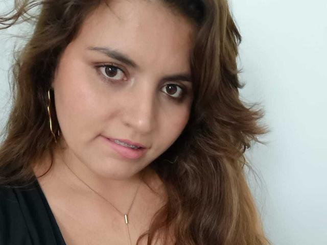 SharaySexy has wavy brown hair, brown eyes, and is wearing gold hoop earrings and a black top with a thin necklace. She's smiling slightly with pink lipstick.