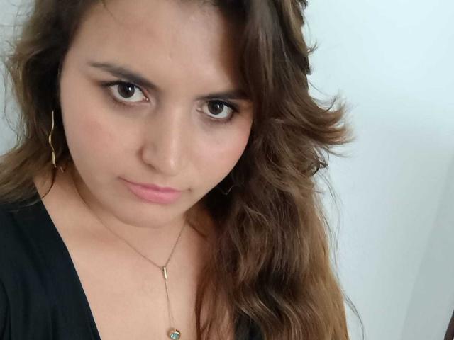 SharaySexy has brown wavy hair, light skin, and brown eyes, wearing a black top and gold earrings, with a slight smirk and a necklace.