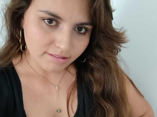 SharaySexy with wavy brown hair, brown eyes, black top, gold hoop earrings, and a gold necklace with a small green pendant, looking slightly down with a subtle smile.