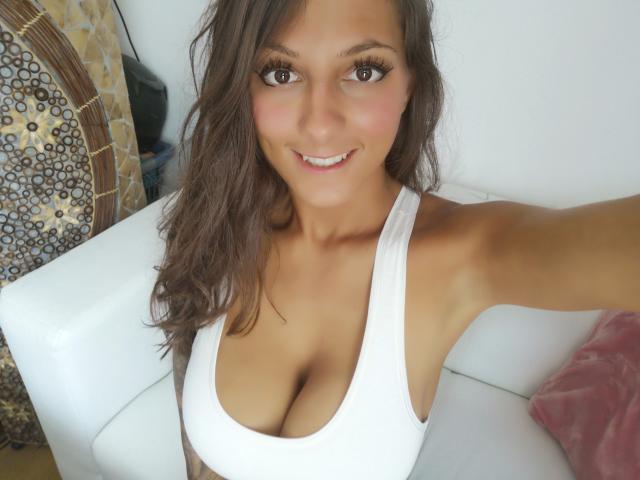 KaitySun-hot with wavy brown hair, large tits in a white tank top, smiling at the camera, sitting on a white couch. She has long eyelashes and a cute face.