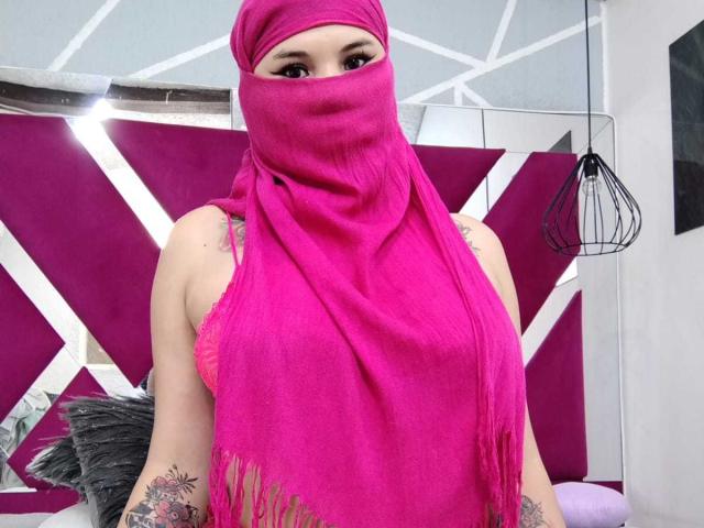 MalakCote portant un foulard rose qui lui couvre la tête et la poitrine, laissant apparaître ses bras tatoués, avec des yeux marron, un foulard rose à franges, et un fond rose-gris..