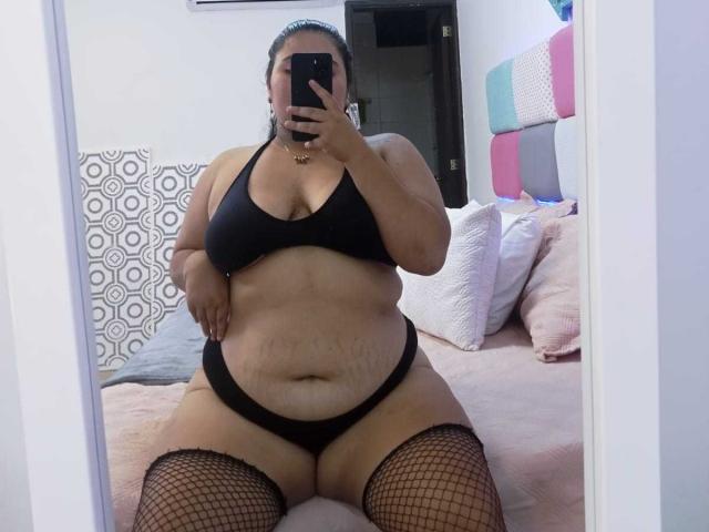 ZoeGiil, plus-sized with large tits and belly, wearing black bra, panties, and fishnet stockings, sitting on bed, taking mirror selfie with phone, covering face.