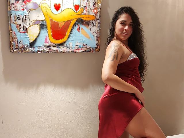 AlejandraMontana in a red satin dress with a white lace trim, showing her ass and a thigh tattoo, standing beside a colorful abstract painting.