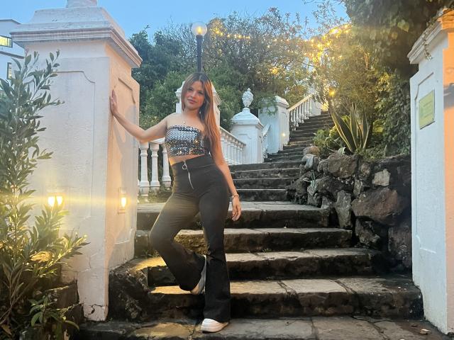 ShantalSummer standing on stone stairs, wearing a sparkly strapless top and black flared pants, with white sneakers, leaning against a white wall, smiling at the camera.