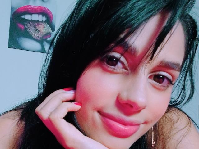 AnaisHilton has dark hair, pink lipstick, and a slight smile. She's touching her cheek with pink nails, showing brown eyes. A photo of red lips with a coin is in the background.
