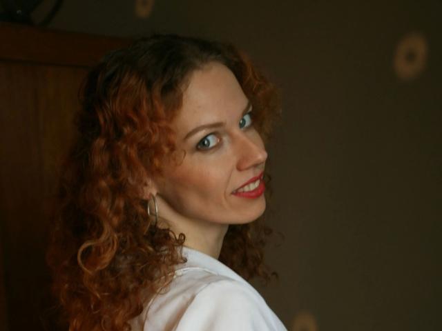 HotRedKinky-hot with curly red hair, blue eyes, red lipstick, smiling at the camera, wearing a white shirt, looking over her shoulder, hoop earrings visible.