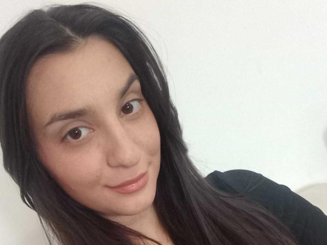 EvaLynda tiene ojos marrón oscuro, cabello negro largo y una sonrisa sutil. Lleva una blusa negra, tiene la piel clara y un aspecto natural y encantador..