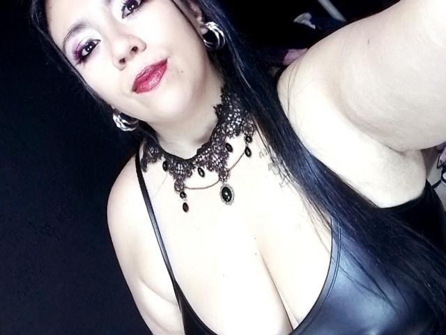LexyDom with black leather top, showing her big tits, wearing a black ornate necklace, and red lipstick, has long black hair, and silver hoop earrings, in a selfie.