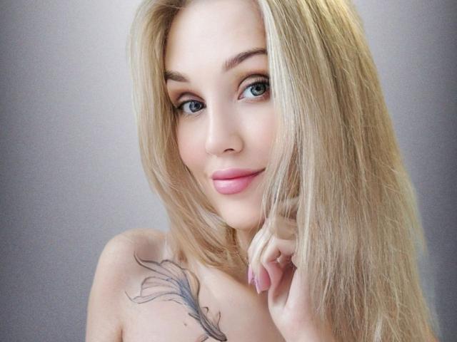 Angelovna with long blonde hair, blue eyes, pink lips, and a feather tattoo on her shoulder, gently touching her chin. She's smiling softly, looking directly at the camera.