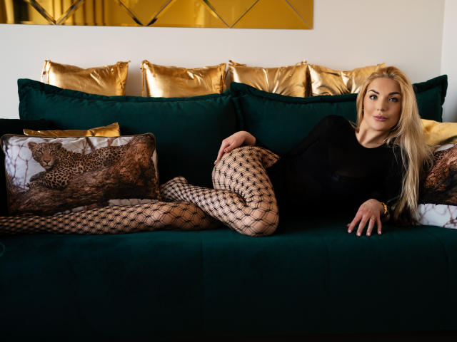 AulloraFlirt, blonde, lying on green velvet couch, wearing black sheer top, fishnet pants, gold pillows, leopard print pillows, sexy, legs crossed, right arm on couch, left arm behind.