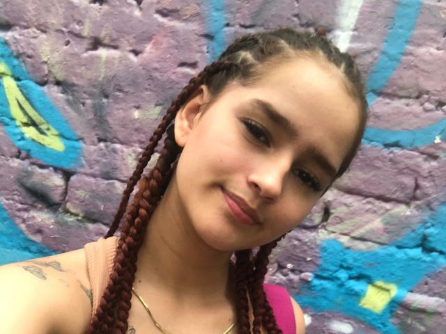Melody Mansson tiene el cabello castaño trenzado, piel clara y pequeños tatuajes en el hombro. Está sonriendo con lápiz labial rosa y lleva una blusa rosa, mostrando sus senos..
