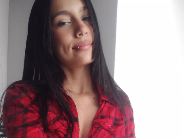KataleyaMansson has medium brown skin, long black hair, and is wearing a red plaid shirt. She's smiling with slightly parted lips and has a small nose piercing.