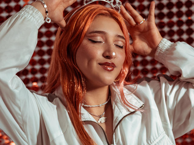 IsabellaClown with orange hair, wearing a white jacket, cat ears, and eyeliner, smiling with eyes closed, hands up, showing off jewelry and a necklace.