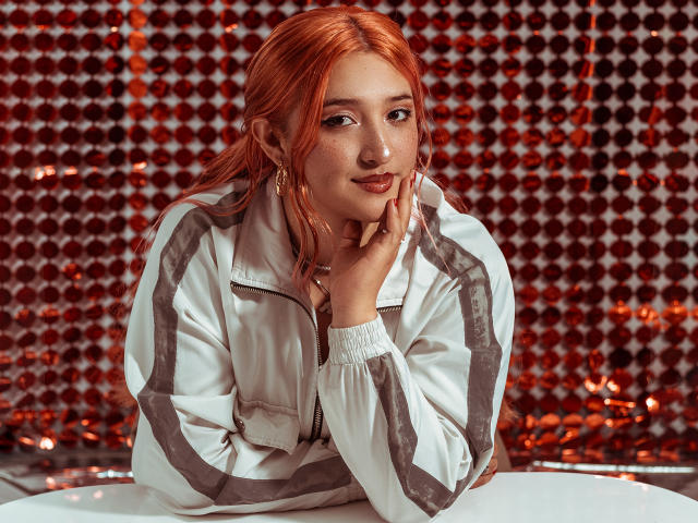 IsabellaClown with red hair, freckles, and hoop earrings, wearing a white track jacket, resting her chin on her hand, smiling softly. She has small tits and a cute face.