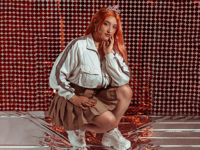 IsabellaClown with orange hair, white jacket, brown skirt, and white sneakers, squatting, hand on chin, red nails, tiara, and a bracelet, in a shiny metallic setting.
