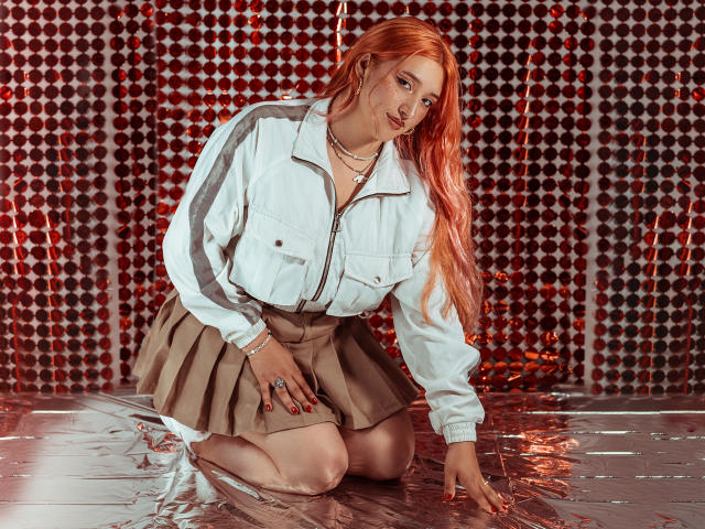 IsabellaClown with red hair, white jacket, and beige skirt, kneeling on shiny red and silver paper, looking at camera, showing her pussy slightly through skirt, in sexy pose.