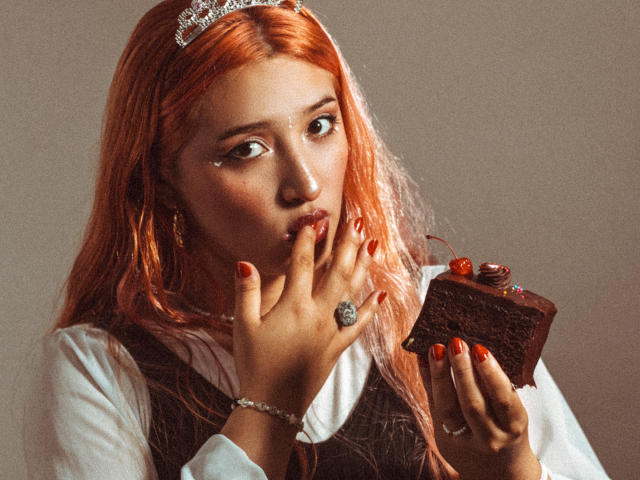 IsabellaClown with orange hair, wearing a tiara, biting chocolate from a dark chocolate slice with red berries, holding it with red nails, a ring, and a bracelet.