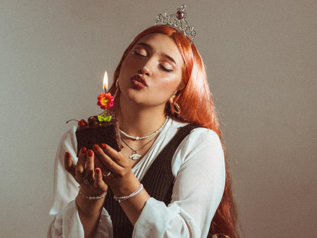 IsabellaClown with red hair, wearing a silver tiara, holding a candlelit chocolate cake with a flower, eyes closed, lipstick on, red nails, white shirt, black vest, necklace.