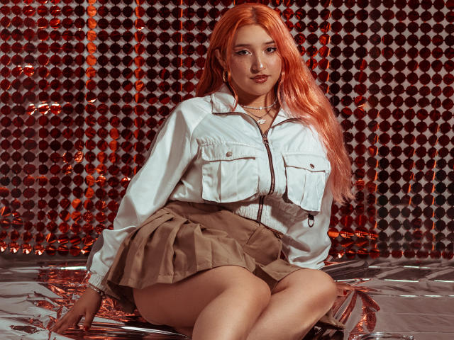 IsabellaClown with long orange hair, white jacket, brown skirt, sitting with legs together, showing her ass, against shiny red-patterned background. She has a cute face and is looking at camera.