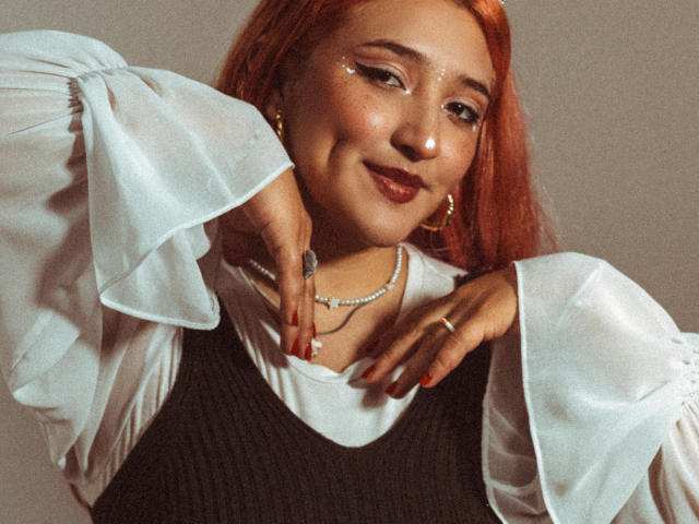 IsabellaClown has red hair, wearing a white blouse with puffy sleeves and a black dress, hands near her face with red nails, and a pearl necklace, smiling.