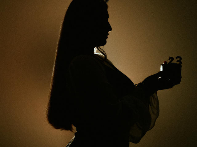 Silhouetted profile of IsabellaClown with long hair, holding a small red light in her right hand, her tits slightly visible, against a dark brown background.