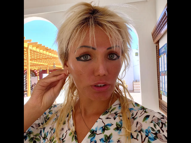 SexyNat-hot with blonde hair, green eyes, and a floral shirt, looking at the camera with a slight pout, hand in hair, and a small freckle on her cheek.