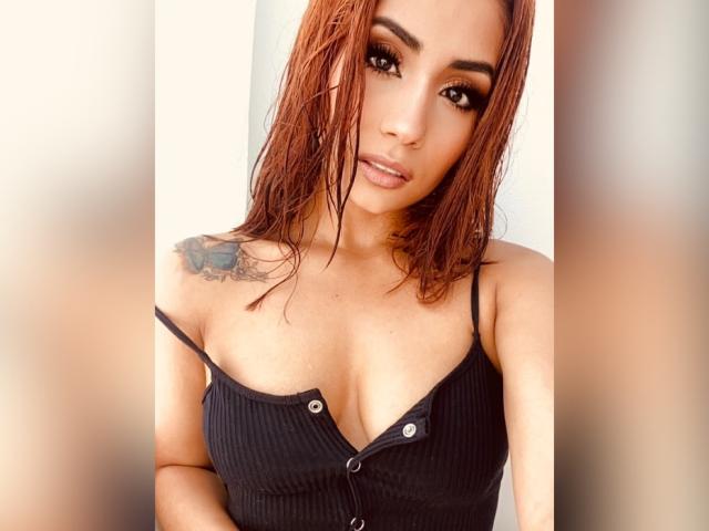 KattyWattson with wet, red hair, wearing a black ribbed tank top, showing her small tits, with a tattoo on her right shoulder, and a sexy, confident expression.
