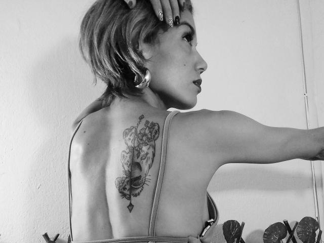 Veroo69 with short hair, back tattoo of a woman with flowers and insects, wearing a strapless top, right arm raised, left hand on head, black-and-white photo.