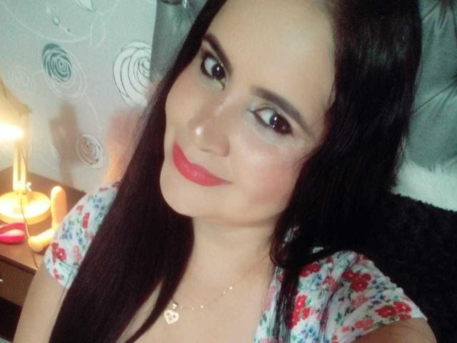 CoralRed, avec ses longs cheveux noirs, son rouge à lèvres rouge et sa chemise à fleurs, sourit à l'appareil photo, montrant son joli visage et ses petites seins mignons. Elle porte un collier en or..