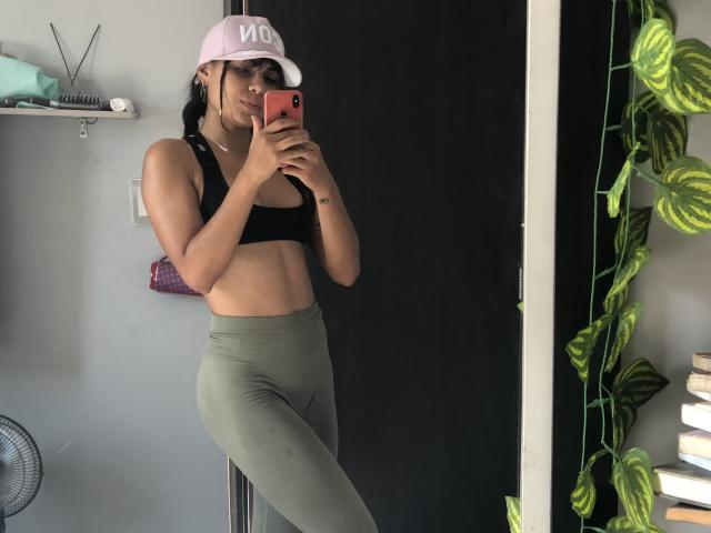 AnnieHal with tan skin, black sports bra showing her tits, green high-waisted leggings, pink hat, taking a mirror selfie with red phone, standing in a room with plants.