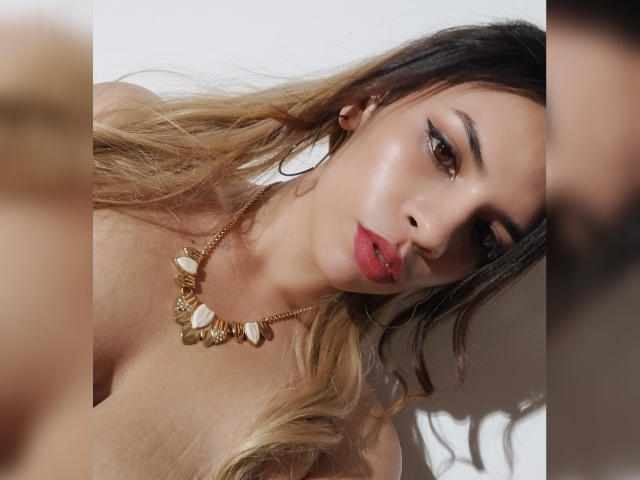 VioletaStone with blonde ombre hair, red lipstick, and gold necklace featuring shells, has shiny skin and is looking down with a serious expression, showing her tits.