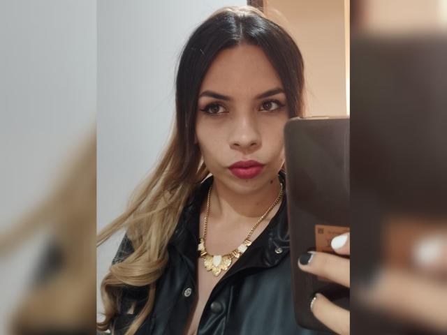 VioletaStone with long ombre hair, wearing a black leather jacket and gold necklace, taking a mirror selfie with pink lipstick and black nail polish.