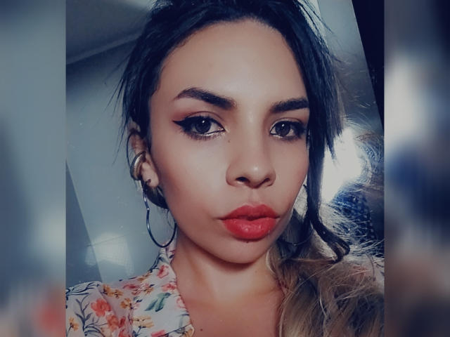 VioletaStone has dark brown hair, red lipstick, and thick eyebrows with winged eyeliner. She's wearing a floral top and large hoop earrings. Her skin is tan and smooth.