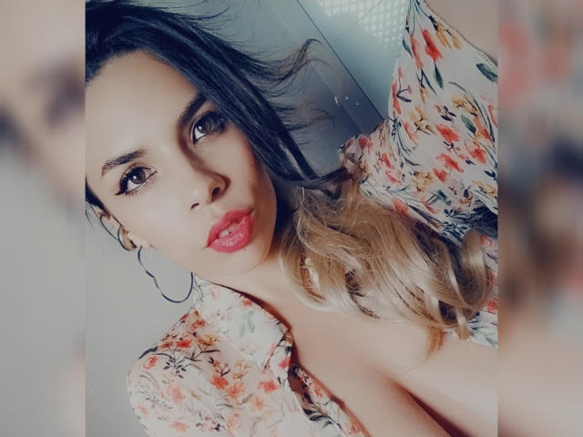 VioletaStone with red lips, brown eyes, and blonde-to-black ombre hair, wearing a floral shirt, showing cleavage, large hoop earrings, and a slight pout.